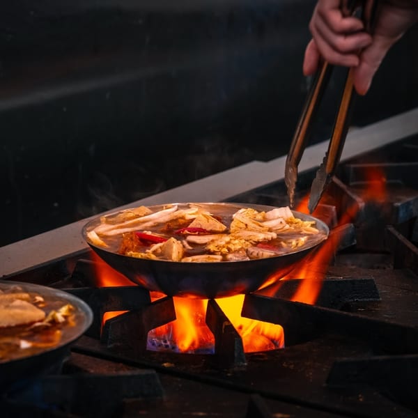 A chef uses tongs to stir a pan of hotpot cooking over an open flame on a stove.