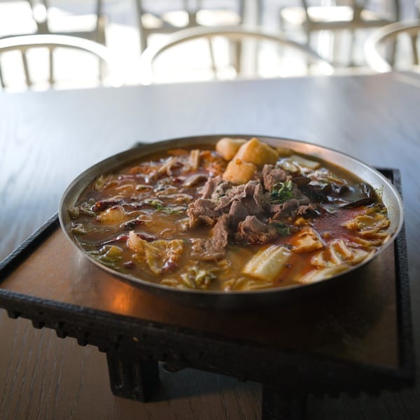 A large metal bowl of spicy hot pot with meat, vegetables, and tofu on a wooden table.