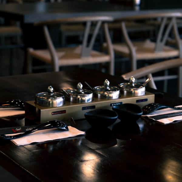 A dark wooden table set with condiment containers, black bowls, and spoons in a restaurant.