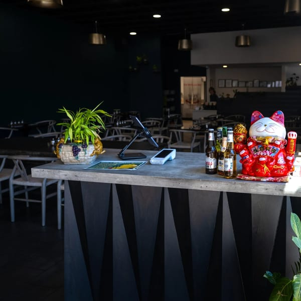 A restaurant's front counter with a lucky cat, beer bottles, a plant, and payment devices.